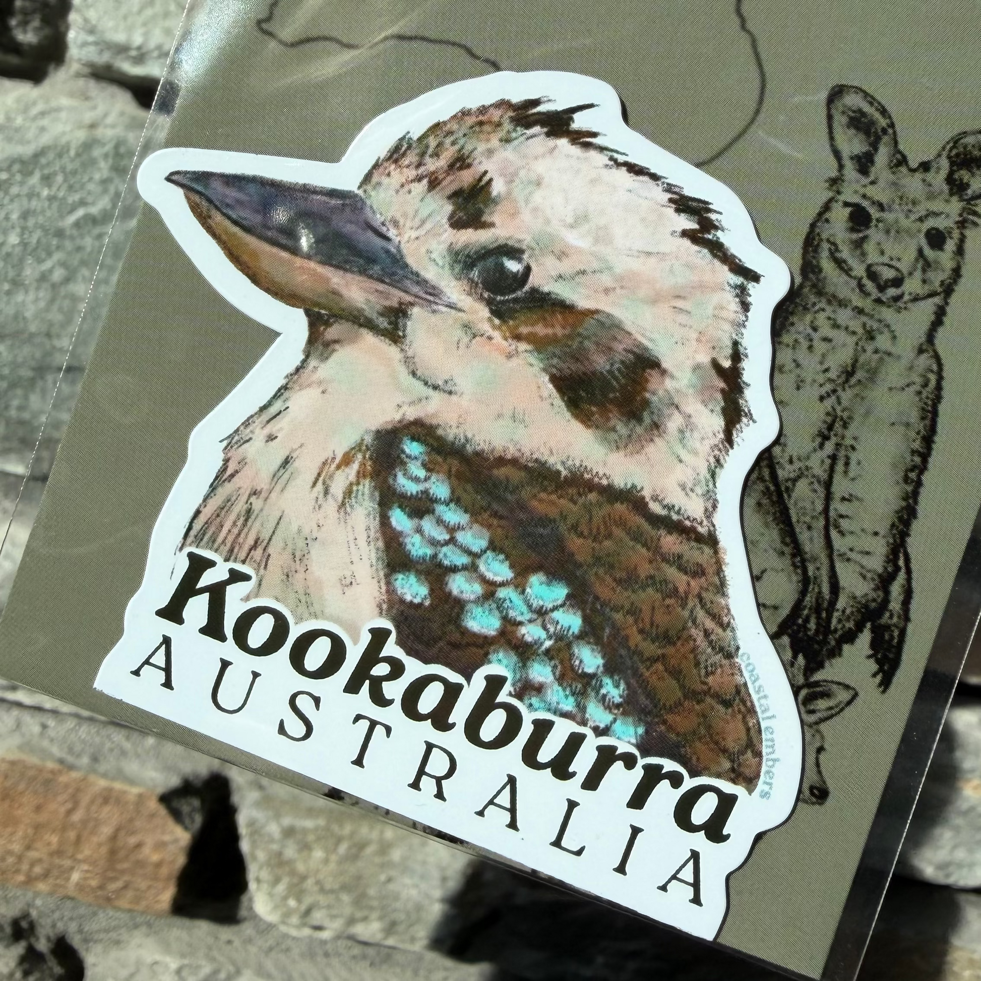 A die-cut sticker featuring a detailed illustration of a kookaburra with the text 'Kookaburra AUSTRALIA' printed below it, attached to a backing card. The backing card also shows a line drawing of a wallaroo and the partial text 'Aussie Mag'.