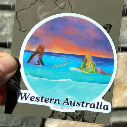 A hand holds a packaged round sticker featuring a colorful Western Australia coastal scene with two mermaids, labeled 'Aussie Magnet' on a gray backing card, against a stone wall background.